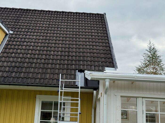 Tak på hus med trapetsformiga pannor möter ett uterums vita tak, stege lutad mot väggen. Brist på avslut gör att taket ser avhugget ut.