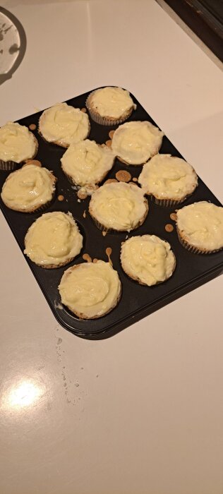 Morotsmuffins med glasyr i muffinsformar på en svart plåt.