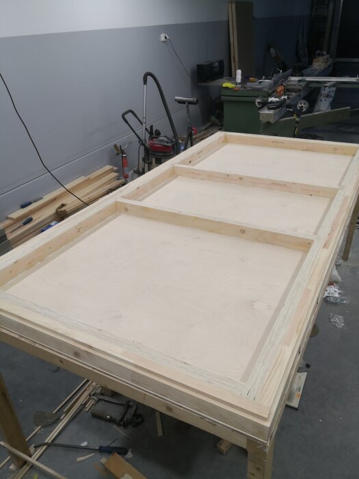 Fingerskarvade dörrar under tillverkning på arbetsbänk med plywood och kertopuu i verkstadsmiljö, omgivet av verktyg och byggmaterial.