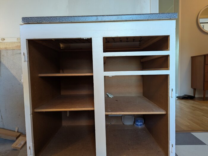 Öppet skåp med hyllor, utan dörrar, i trä med vitmålad ram och grå bänkskiva. Skåpet är under renovering med tillbehör runtom.