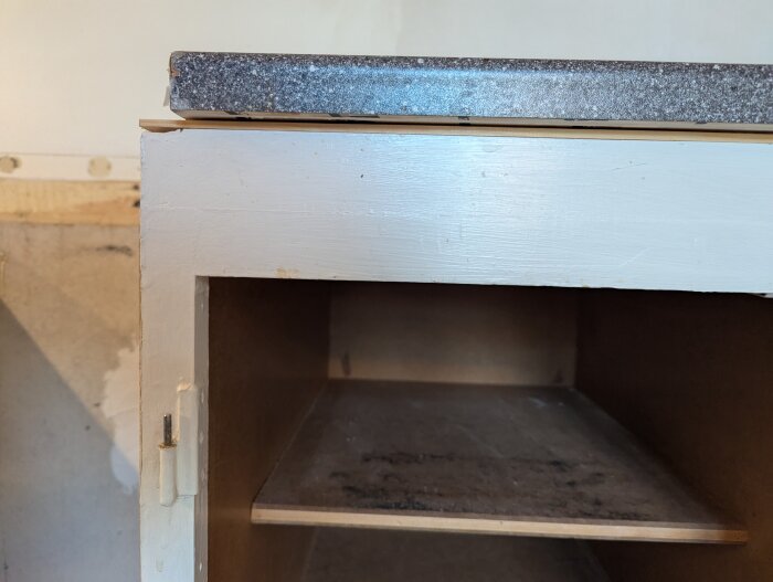 Närbild av en vit skåpram med en tunn trälist på toppen under en bänkskiva. Skåpets insida och hylla är delvis synliga.