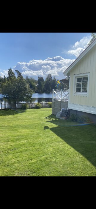 Vit träfasad med fönster, svensk flagga på veranda, utsikt över gräsmatta och sjö med skog och molnig himmel i bakgrunden.