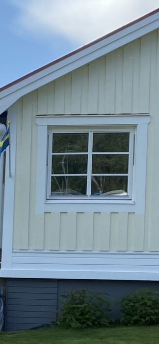 Fönster med vita fönsterfoder på en gul fasad, med växtlighet nedanför och en svensk flagga till vänster.