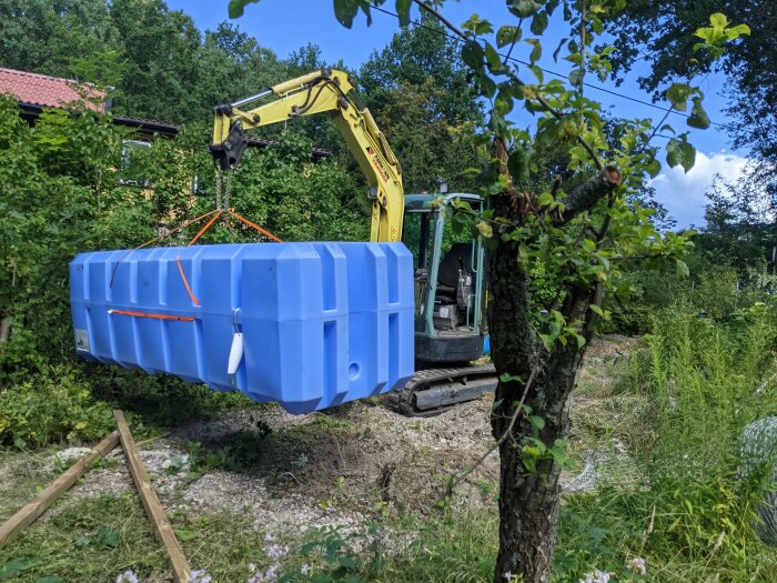 En grävmaskin lyfter en stor blå plastbehållare i en trädgård omgiven av grönska.