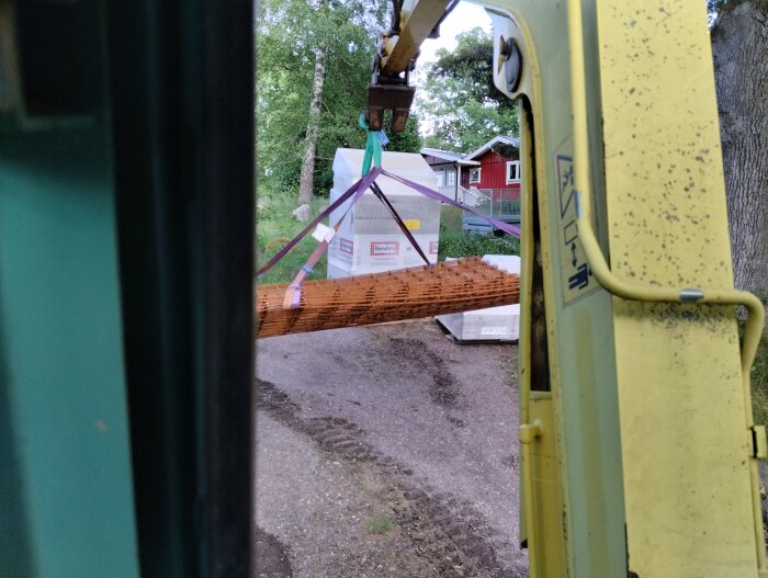 En gul grävare lyfter en stor last med brunt byggmaterial med hjälp av stroppar, på en byggarbetsplats med synliga träd och hus i bakgrunden.