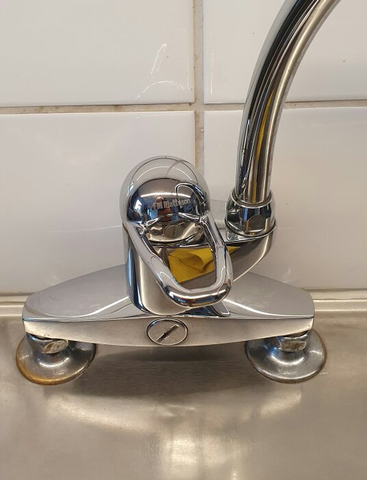 En kromad FM Mattson kökskran med en gul märkning, installerad ovanpå en rostfri diskbänk mot en kaklad vägg.