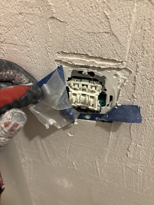 En person håller i en delvis bortmonterad strömbrytare på en vägg med vit tejp runt om.