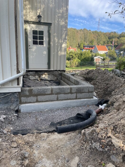 Husgrund under arbete med dräneringsrör och betongblock, vid sidan av ett äldre hus. Trädgård och hus i bakgrunden.