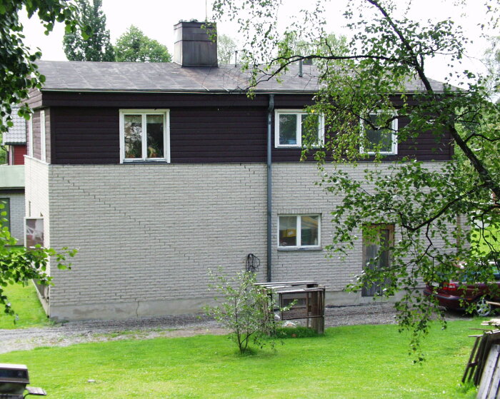 Tvåvåningshus med mörk stående panel och ljus tegelvägg, omgiven av träd och grönska.