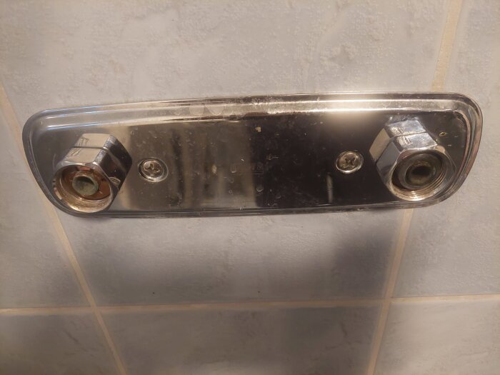 Äldre duschblandare med täckplatta mellan rören på vägg, med synliga muttrar och fästen, i ett badrum.