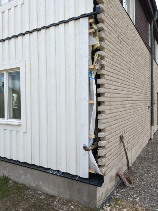 Stående vit panel ersätter gammal fasad på hus från 1912, delvis rivet för att visa materiallagren och byggprocessen. Verktyg syns vid sidan.