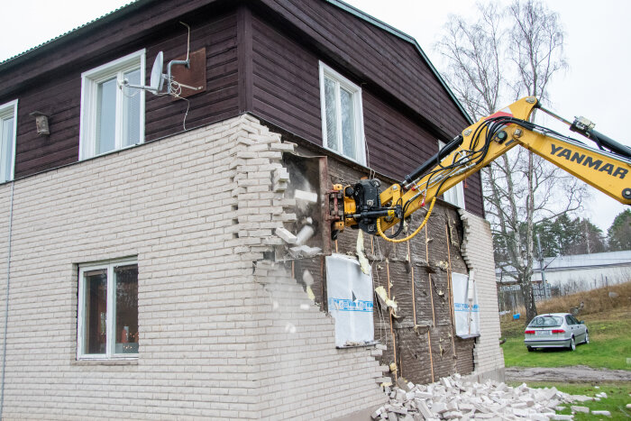Fasadrenovering på ett hus från 1912 där en grävmaskin river bort gammalt tegel, exponerar träpanel och isolering.