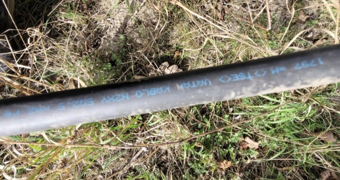 Svart elkabel med tryckt text i blått ligger på marken med gräs och löv runtomkring.