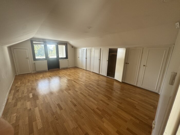 Ett stort rum med trägolv och inbyggda garderober, med fönster och vitmålade väggar.