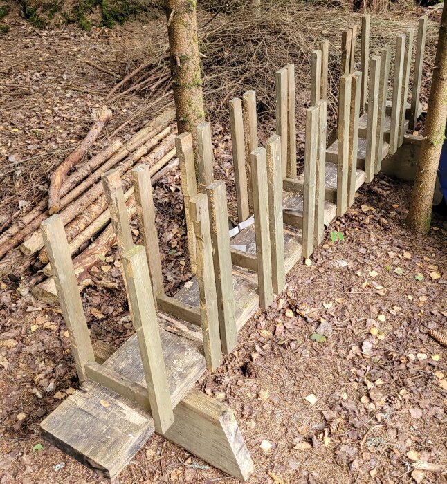 Träbock med vertikala trästöd, placerad i en skogsmiljö, omgiven av träd och grenar på marken. Används för att såga upp ved.