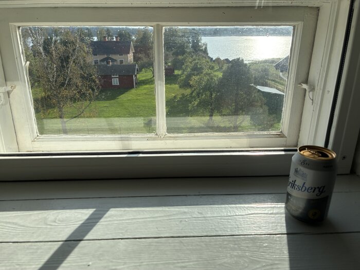 Utsikt genom fönster med en burk Eriksberg-öl på fönsterbrädan, trädgård och sjö syns utanför.