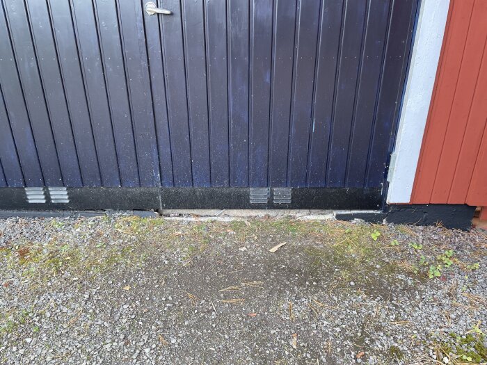 Blå garagedörr med spricka mellan betongplattan och det yttre skiktet, synlig i botten av dörren med grus och gräs framför.