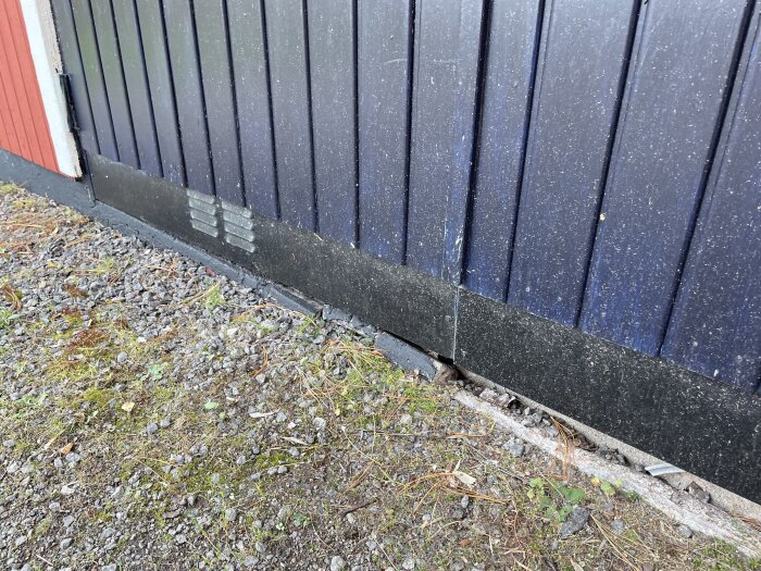 Garagedörr med sprucken bas mellan platta och yttre skikt, som blottar skiktmaterial, med grönt grus och mark med mossa.