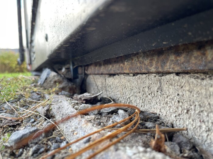 Garagedörr med spricka vid fundamentet, mellan betongplatta och yttre skikt. Rosten syns på metallen, omgivet av stenar och gräsväxt.