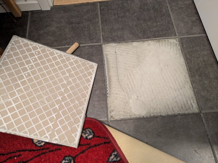 Lös klinkerplatta på hallgolv, borttagen för att visa sprucken fog och saknat fix. Mattkant syns på bilden.