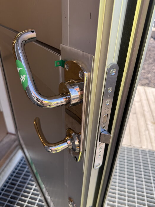 Närbild på ett dörrhandtag och säkerhetslås med grön panikhandtagsmarkering på en metallfärgad dörr.