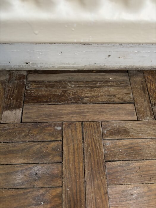 Gamla stavparkettgolv med en slipad planka synlig, mörkare än de omgivande. Golvet visar tecken på tidigare fuktskada.