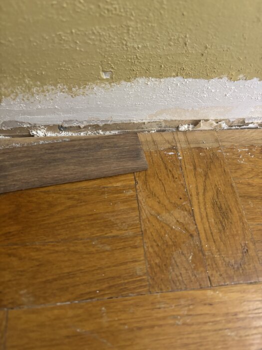 Gammal stavparkett synlig under borttaget klickgolv, närbild av mörka träplankor vid en vägg med borttagen golvlist och synliga skador.