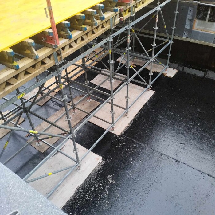 Byggställning med träbjälkar och svarttätad yta.