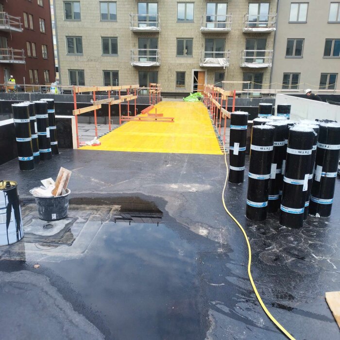 Byggarbetsplats med asfalt, rullar av takpapp och en gul ramp. Bakom syns ett flerfamiljshus under konstruktion.