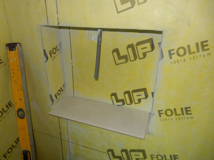 Badrumsvägg med tätskikt av gul folie, syns hål för hylla och rör, samt en gul vattenpass i hörnet.