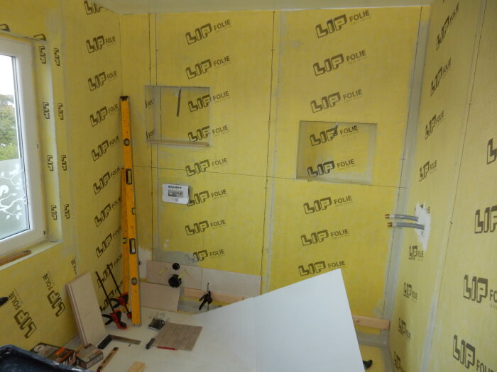 Badrum med tätskikt monterat, skyddspapp med logotyp synlig på väggarna, verktyg och byggmaterial på golvet, fönster till vänster.