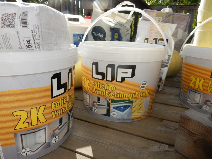 Hinkar med LIP 2K folielim och vattentätningsmaterial placerade på en träyta, omgärdade av renoveringsprodukter för badrum.