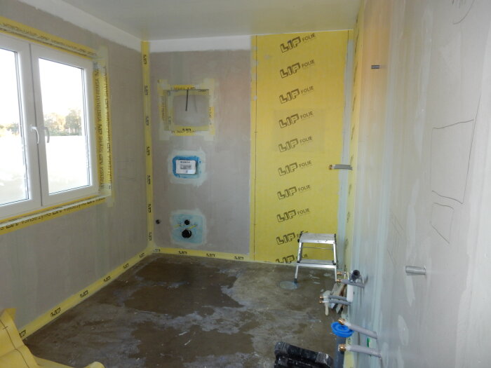 Badrum under renovering med tätskikt på väggarna, rörinstallationer synliga och stege i rummet. Fönster på vänstra väggen.