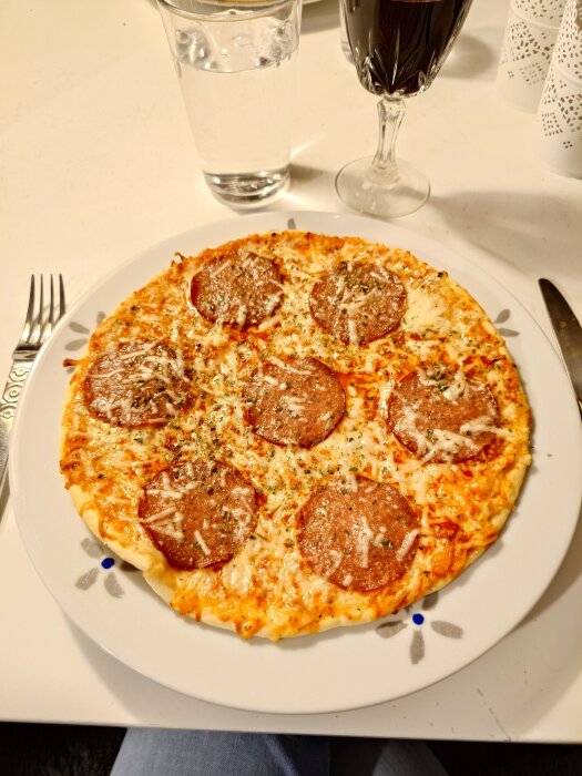 En färdiglagad pepperonipizza på en vit tallrik, serverad med ett glas vatten och ett glas rödvin på ett vitt bord.
