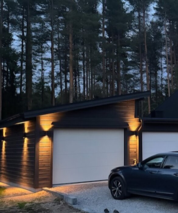 Nybyggt garage med 14 graders pulpettak och liggande träpanel, belyst och omgivet av träd. Svart bil parkerad framför på grusad uppfart.