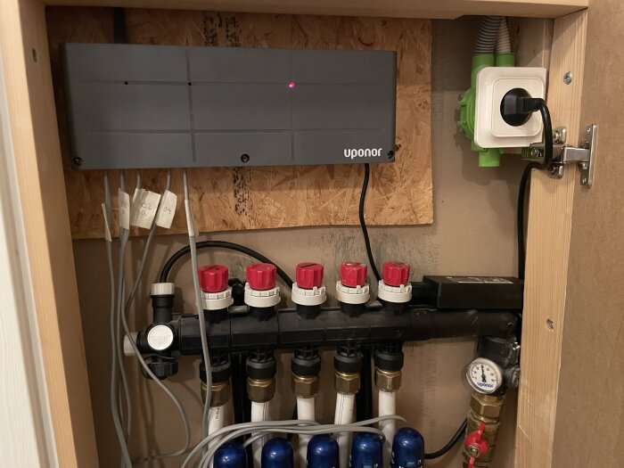 Elcentral och rördragning i ett skåp, med kopplingsbox märkt "Uponor", kablar och röranslutningar synliga. En nätadapter inkopplad.