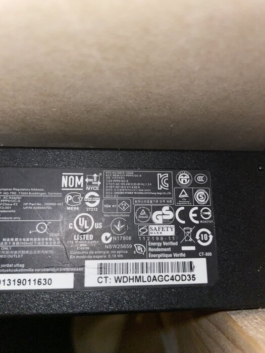 Etikett på en datorladdare med olika certifieringssymboler, specifikationer och en streckkod.