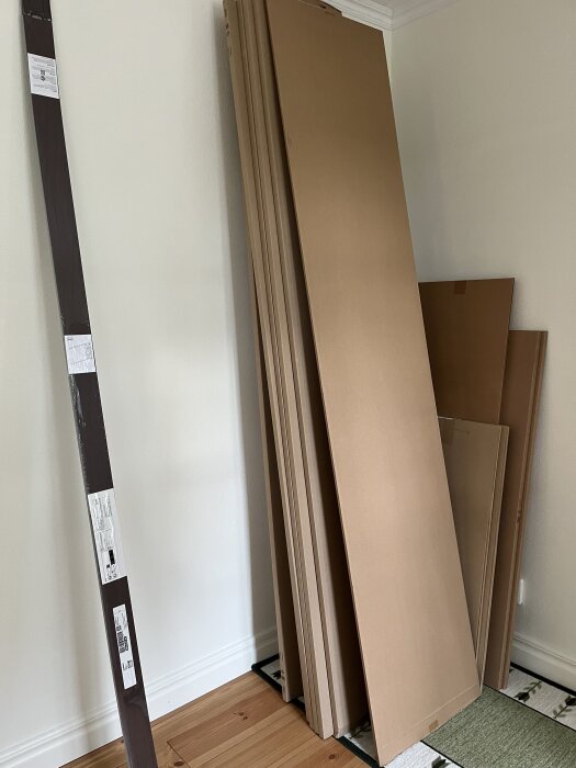Staplade platta kartonger med möbelpaket från IKEA, står lutade mot en vägg på ett trägolv och matta.