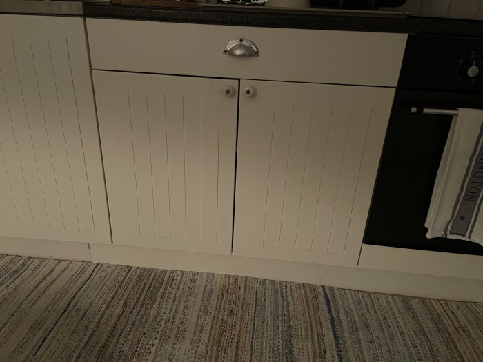 Vitt köksskåp med Ståt luckor och en handduk hänger på ugnshandtaget. Diskmaskin önskas installeras genom anpassning av skåpen.