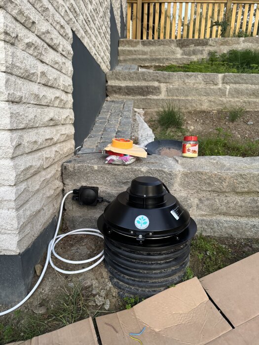 Svart ventilationsfläkt installerad utomhus mot en murad trappa, med en vit kabel och olika verktyg liggandes bredvid.