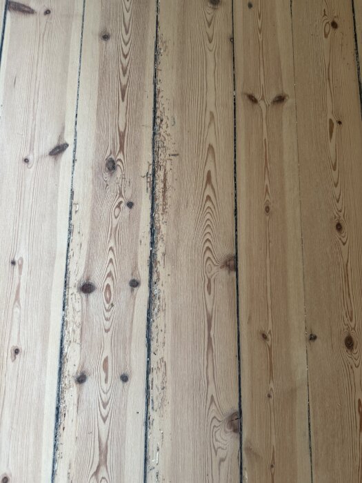 Ett gammalt furugolv med synliga märken och ojämn slipning; vissa delar har börjat tappa hårdvaxolja.