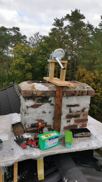 Brick chimney with winch setup and tools, including a drill and gloves, placed on a covered rooftop in a wooded area.