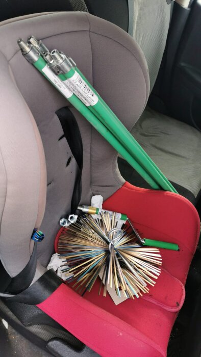 Gröna teleskopstänger och en cirkulär borste med metallborst ligger på ett rött bilbarnstolssäte, troligen för sotrengöring.