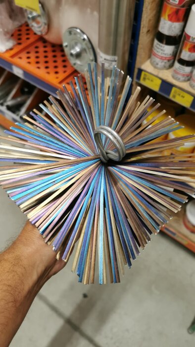 Används i en renoveringsprocess, bilden visar en hand som håller i en chimneysotborste med färgglada och metalliska borststrån, fotograferad i en butikshylla.