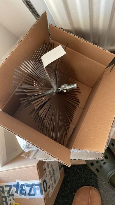 A chimney cleaning brush with metal bristles is shown inside a cardboard box, part of tools for a renovation project.