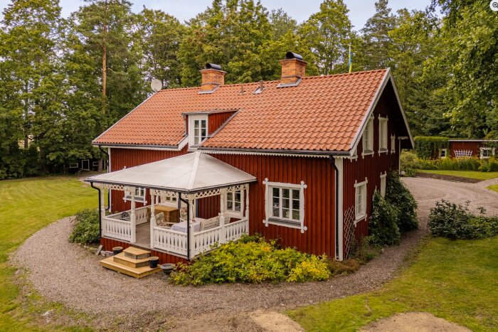 Rött timmerhus med vit veranda, brunt tegeltak och takkupa. Omgiven av trädgård och grusgång, med skog i bakgrunden.
