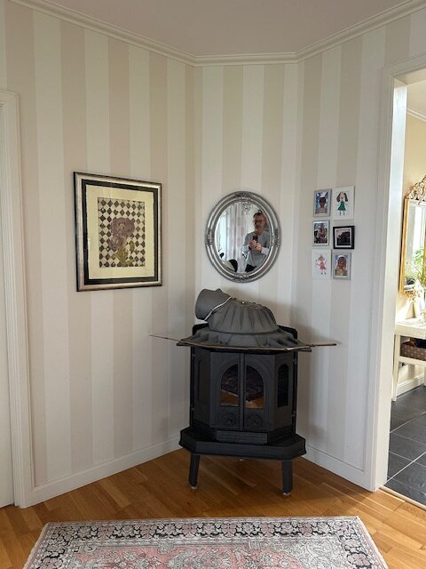 Hörn av rum med vit vägg, rund spegel, braskamin, konstverk och familjefoton på väggen, samt en matta på golvet.