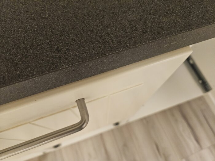 Närbild på en mörk köksbänkskiva monterad ovanpå ett vitt köksskåp med ett metallhandtag.