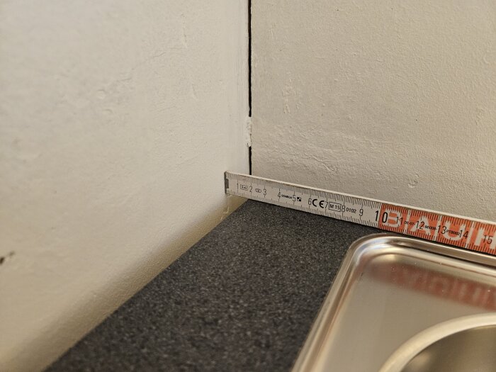 Måttband visar avstånd mellan köksbänk och vägg, cirka 20 mm, i ett kök med en svart bänkskiva och diskho av rostfritt stål.