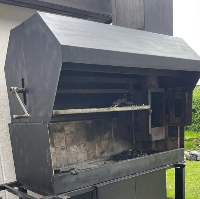 Svart trädgårdsgrill med höj- och sänkbart galler, delvis stängt lock och tegelplattor inuti, placerad utomhus.
